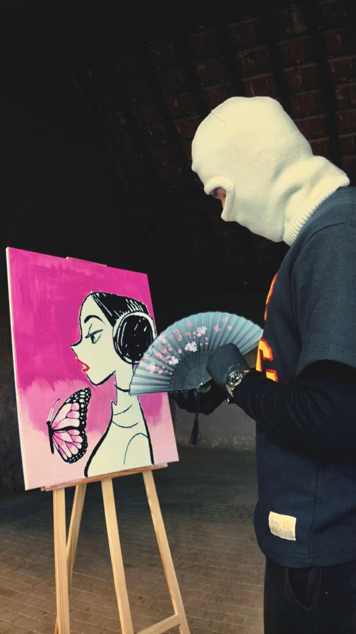 Masked figure standing beside a pink painting with a fan